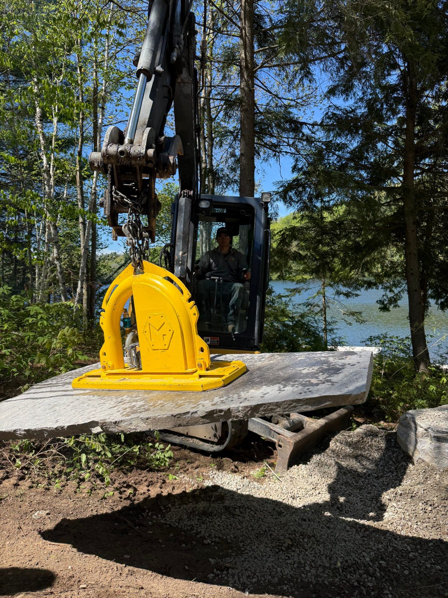Excavator lifting large stone slab in wooded area.