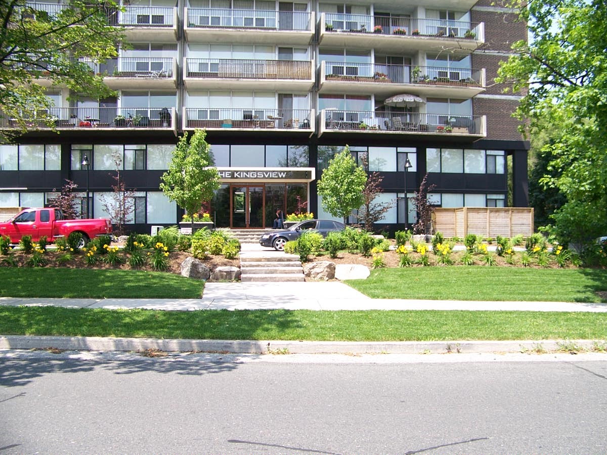Apartment building entrance with landscaping, parked cars.